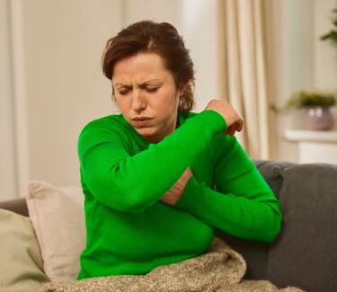 Eine Frau mittleren Alters in einem grünen Pullover sitzt auf einem Sofa und hustet in ihre Armbeuge, ein häufiges Symptom einer Erkältung oder Bronchitis.