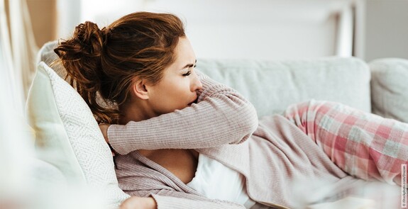 Eine Frau mit Erkältung liegt hustend auf dem Sofa.