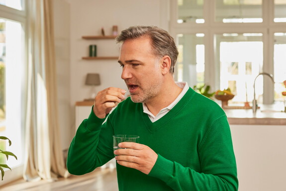 Mann mittleren Alters in grünem Pullover steht in einem hellen Raum und nimmt eine Gelomyrtol Forte Kapsel mit einem Glas Wasser ein.