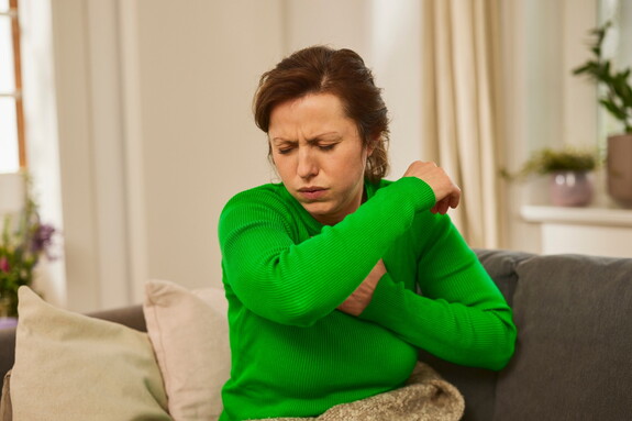 Eine Frau mittleren Alters in einem grünen Pullover sitzt auf einem Sofa und hustet in ihre Armbeuge.