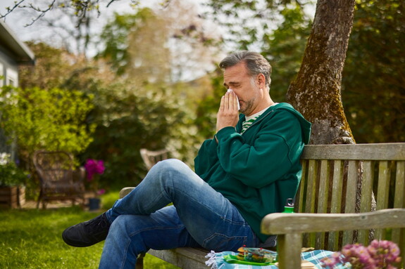 Mann in grünem Pullover sitzt auf einer Gartenbank und putzt sich die Nase mit einem Taschentuch.