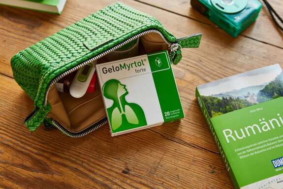 Eine geöffnete grüne Reisetasche auf einem Holztisch, die eine Packung Gelomyrtol Forte und andere Reiseutensilien enthält, liegt bereit für die Gesundheitsvorsorge unterwegs.