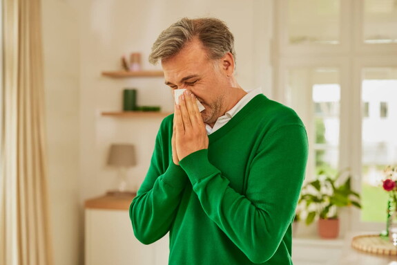 Älterer Herr in grünem Pullover hält sich in einem hellen Raum ein Taschentuch vor das Gesicht, während er niest, was auf Erkältungssymptome hinweist.