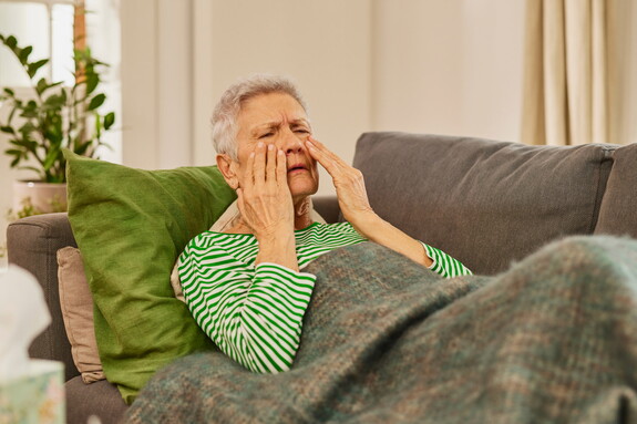 Eine ältere Frau mit grauen Haaren, gekleidet in einem gestreiften grün-weißen Pullover, sitzt auf einem Sofa, hält sich die Handflächen an die Nebenhöhlen und scheint Schmerzen zu empfinden, möglicherweise aufgrund einer Kieferhöhlenentzündung.