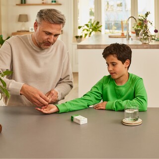 Erwachsener Mann reicht einem Jungen in einem grünen Pullover eine Kapsel Gelomyrtol Forte am Küchentisch als Teil der Gesundheitspflege.