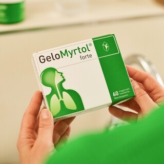 Person in grünem Pullover hält eine Packung Gelomyrtol Forte im Badezimmer, reflektiert in einem Spiegel, was auf die tägliche Routine der Gesundheitsvorsorge hinweist.