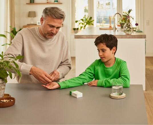 Erwachsener Mann reicht einem Jungen in einem grünen Pullover eine Kapsel Gelomyrtol Forte am Küchentisch als Teil der Gesundheitspflege.