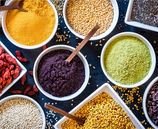 Verschiedene bunte Superfood-Pulver in Schalen, darunter gelbes, grünes und violettes Pulver, arrangiert auf einer dunklen Oberfläche, umgeben von Samen und getrockneten Früchten.