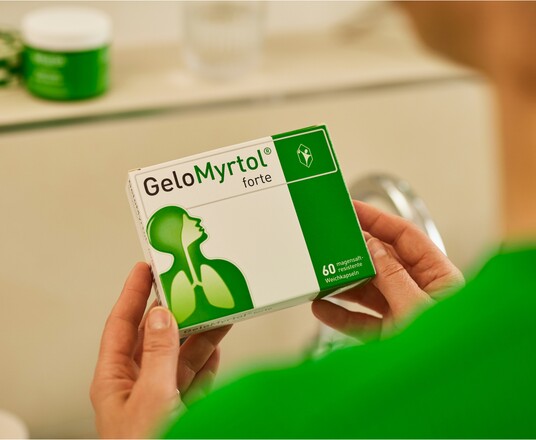 Hände halten eine Packung GeloMyrtol forte, die im Vordergrund scharf zu sehen ist, mit dem Logo und der grünen Markenfarbe deutlich sichtbar, im Hintergrund unscharf eine weitere Produktverpackung.