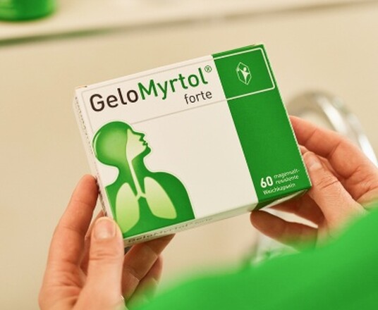 Person in grünem Pullover hält eine Packung Gelomyrtol Forte im Badezimmer, reflektiert in einem Spiegel, was auf die tägliche Routine der Gesundheitsvorsorge hinweist.