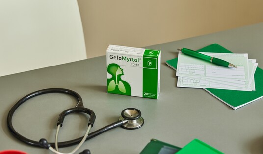 Auf einem Schreibtisch liegt eine geöffnete Packung Gelomyrtol Forte neben medizinischen Utensilien, darunter ein Stethoskop und ein Notizblock.