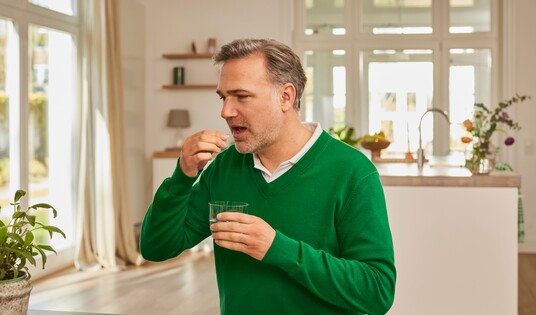 Mann mittleren Alters in grünem Pullover steht in einem hellen Raum und nimmt eine Gelomyrtol Forte Kapsel mit einem Glas Wasser ein.