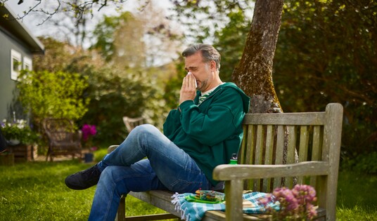 Mann in grünem Pullover sitzt auf einer Gartenbank und putzt sich die Nase mit einem Taschentuch.