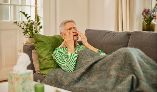 Eine ältere Frau mit grauen Haaren, gekleidet in einem gestreiften grün-weißen Pullover, sitzt auf einem Sofa, hält sich die Handflächen an die Nebenhöhlen und scheint Schmerzen zu empfinden, möglicherweise aufgrund einer Kieferhöhlenentzündung.