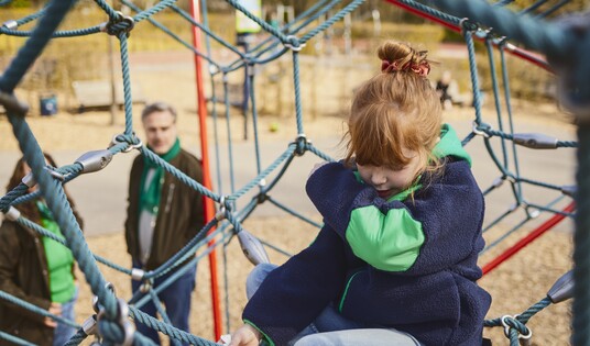 Kind mit Husten auf dem Spielplatz, die Eltern stehen im Hintergrund.