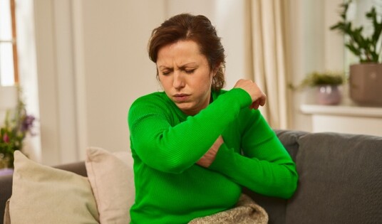 Eine Frau in einem grünen Pullover sitzt auf einem Sofa und hustet in ihren Ellenbogen.