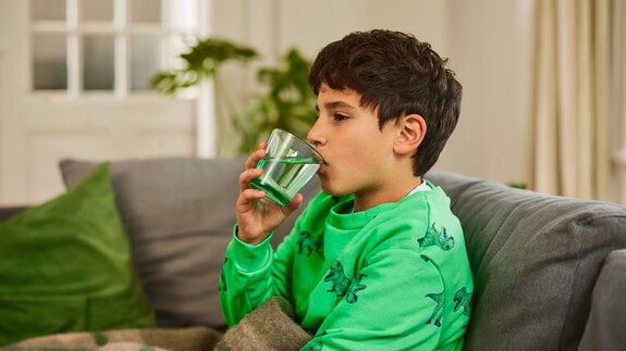 Kind mit Erkältung trinkt ein Glas Wasser auf dem Sofa.