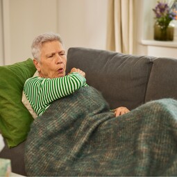 Gelomyrtol Forte - Effektive Unterstützung bei Sinusitis und Kieferhöhlenentzündung. Eine ältere Dame mit grauen Haaren in einem grün-weiß gestreiften Pullover sitzt auf einem Sofa und hustet in ihre Armbeuge.