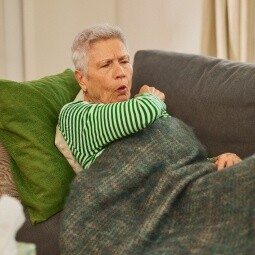 Gelomyrtol Forte - Effektive Unterstützung bei Sinusitis und Kieferhöhlenentzündung. Eine ältere Dame mit grauen Haaren in einem grün-weiß gestreiften Pullover sitzt auf einem Sofa und hustet in ihre Armbeuge.