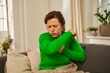 Eine Frau mittleren Alters in einem grünen Pullover sitzt auf einem Sofa und hustet in ihre Armbeuge.