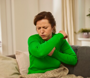 Eine Frau mittleren Alters in einem grünen Pullover sitzt auf einem Sofa und hustet in ihre Armbeuge.