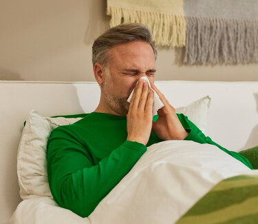 Mann mittleren Alters in grünem Pullover liegt im Bett und putzt sich die Nase mit einem Taschentuch, was auf Erkältungssymptome wie Schnupfen hinweist.