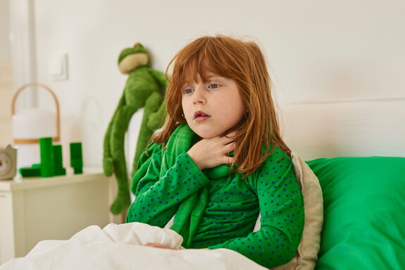 Ein junges Mädchen mit roten Haaren und einem grünen Tupfenpullover sitzt auf einem Bett, hält sich den Hals und schaut besorgt, was auf Halsschmerzen oder eine beginnende Bronchitis hindeuten könnte.