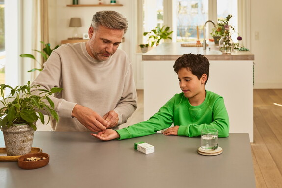 Erwachsener Mann reicht einem Jungen in einem grünen Pullover eine Kapsel Gelomyrtol Forte am Küchentisch als Teil der Gesundheitspflege.