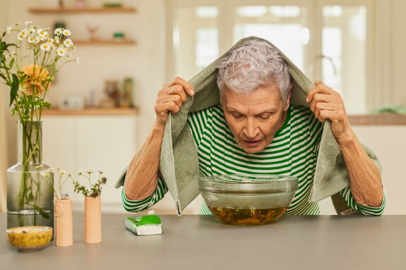 Ältere Frau mit einem Handtuch über dem Kopf beugt sich über eine Schüssel mit dampfendem Wasser zur Linderung von Erkältungssymptomen.