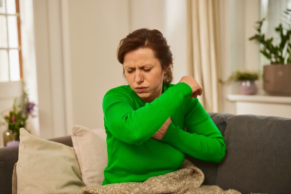 Gelomyrtol Forte - Effektive Linderung bei Erkältungshusten. Eine Frau mittleren Alters in einem grünen Pullover sitzt auf einem Sofa und hustet in ihre Armbeuge.