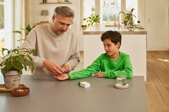 Unterstützung der Familie bei der Gesundheitspflege - Gemeinsame Fürsorge von Generation zu Generation. Erwachsener Mann reicht einem Jungen in einem grünen Pullover eine Kapsel Gelomyrtol Forte am Küchentisch als Teil der Gesundheitspflege.