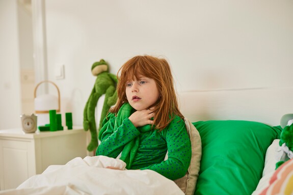 Ein junges Mädchen mit roten Haaren und einem grünen Tupfenpullover sitzt auf einem Bett, hält sich den Hals und schaut besorgt, was auf Halsschmerzen oder eine beginnende Bronchitis hindeuten könnte.