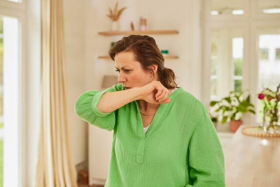 Frau hustet in den Arm – Hinweis auf Husten und Erkältungssymptome bei chronischer Bronchitis. Frau mit grünem Oberteil hustet in ihren Arm in einem hellen, wohnlichen Raum. Symbolisiert Symptome von Bronchitis oder Erkältung.