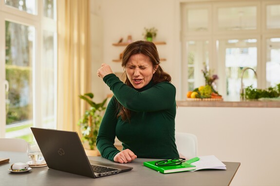 Gelomyrtol Forte – Produktiv arbeiten, auch wenn eine Erkältung zuschlägt. Frau in einem dunkelgrünen Pullover sitzt mit Laptop und Notizblock Zuhause an einem Schreibtisch und hustet in ihre Armbeuge.