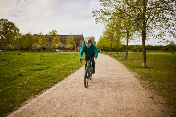 Fahrradfahrer in einem Park – Bewegung an der frischen Luft zur Unterstützung der Atemwege. Mann auf einem Fahrrad fährt auf einem schmalen Schotterweg in einem grünen Park, umgeben von blühenden Bäumen, symbolisch für Aktivität und Atemwegsgesundheit.