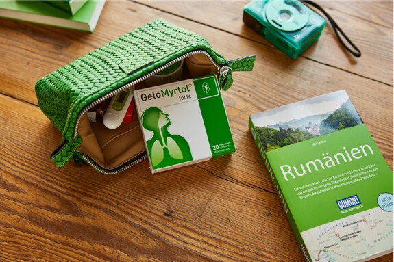 Eine geöffnete grüne Reisetasche auf einem Holztisch, die eine Packung Gelomyrtol Forte und andere Reiseutensilien enthält, liegt bereit für die Gesundheitsvorsorge unterwegs.