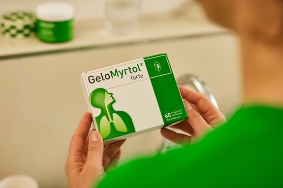 Gelomyrtol Forte – Vertrauen Sie auf bewährte Qualität für Ihre Erkältungslinderung. Hände halten eine Packung GeloMyrtol forte, die im Vordergrund scharf zu sehen ist, mit dem Logo und der grünen Markenfarbe deutlich sichtbar, im Hintergrund unscharf eine weitere Produktverpackung.
