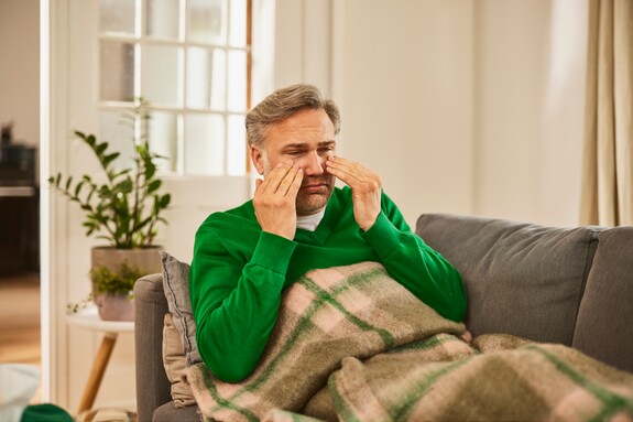 Gelomyrtol Forte – Effektive Linderung der Symptome bei Sinusitis und Förderung des Wohlbefindens zu Hause. Ein älterer Mann mit grauem Haar, in einem grünen Pullover, sitzt auf einem Sofa mit einer Decke über den Beinen und massiert die Bereiche um seine Augen, ein Zeichen für Beschwerden im Bereich der Nasennebenhöhlen.