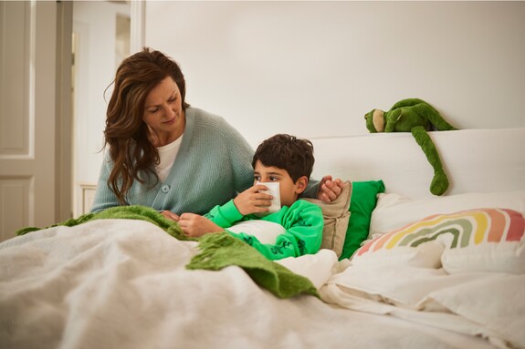 Kind liegt mit Erkältung naseputzend im Bett, die Mutter sitzt daneben.