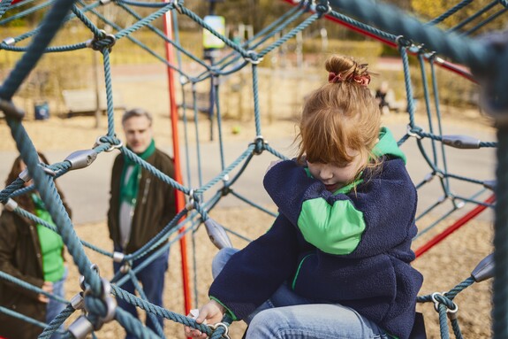 Kind mit Husten auf dem Spielplatz, die Eltern stehen im Hintergrund.