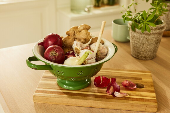Grüne Schale auf einem Holzschneidebrett mit Zutaten für eine gesunde Ernährung, darunter rote Zwiebeln, Knoblauch und Ingwer.