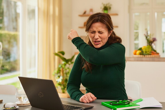 Frau in einem dunkelgrünen Pullover sitzt mit Laptop und Notizblock Zuhause an einem Schreibtisch und hustet in ihre Armbeuge.
