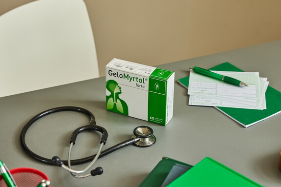 Auf einem Schreibtisch liegt eine geöffnete Packung Gelomyrtol Forte neben medizinischen Utensilien, darunter ein Stethoskop und ein Notizblock.