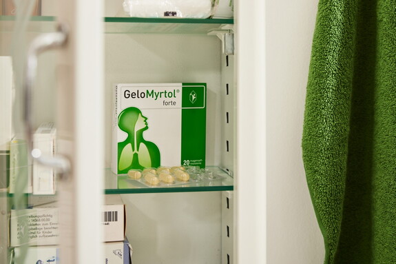 Blick in einen offenen Medizinschrank mit einer Packung Gelomyrtol Forte sichtbar auf einem Regal, neben anderen Gesundheitsprodukten und einem grünen Handtuch an der Tür hängend.