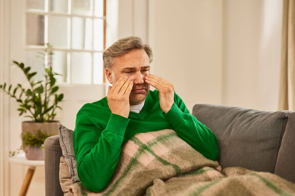 Ein älterer Mann mit grauem Haar, in einem grünen Pullover, sitzt auf einem Sofa mit einer Decke über den Beinen und massiert die Bereiche um seine Augen, ein Zeichen für Beschwerden im Bereich der Nasennebenhöhlen.