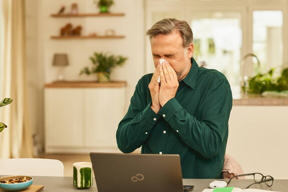 Mann mittleren Alters in einem dunkelgrünen Hemd niest in ein Taschentuch vor einem Laptop auf einem Schreibtisch.