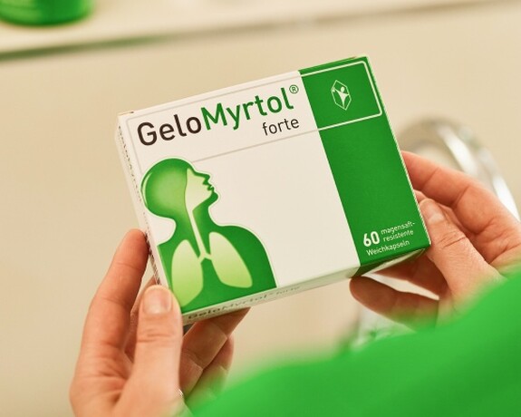 Gelomyrtol Forte – Vertrauen Sie auf bewährte Qualität für Ihre Erkältungslinderung. Person in grünem Pullover hält eine Packung Gelomyrtol Forte im Badezimmer, reflektiert in einem Spiegel, was auf die tägliche Routine der Gesundheitsvorsorge hinweist.