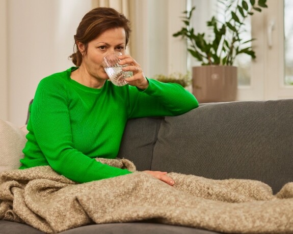 Frau entspannt auf dem Sofa mit einem Glas Wasser - ausreichende Flüssigkeitszufuhr und Ruhe bei Infekten. Frau im grünen Pullover sitzt auf einem Sofa, eingewickelt in eine Decke und trinkt ein Glas Wasser.