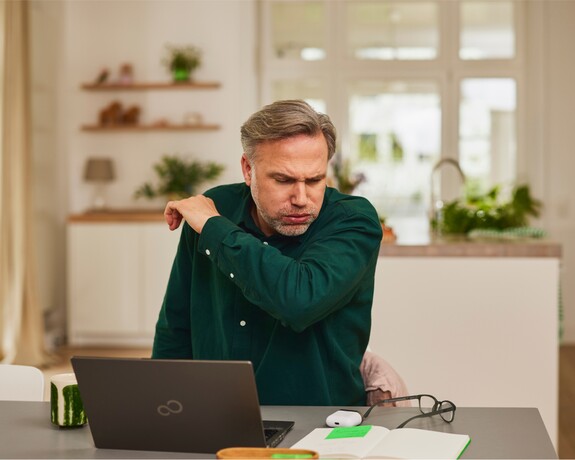 Gelomyrtol Forte – Gesundheit am Arbeitsplatz wahren durch Unterstützung bei Erkältungssymptomen. Mann mittleren Alters in einem dunkelgrünen Pullover hustet in seinen Arm während er Zuhause an einem Schreibtisch mit Laptop sitzt.