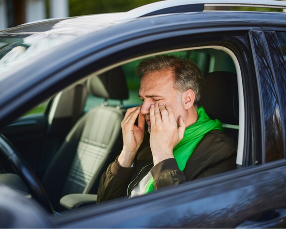Gelomyrtol Forte – Effektive Linderung von Erkältungssymptomen, selbst wenn Sie unterwegs sind. Mann mittleren Alters mit grünem Schal sitzt im Fahrersitz eines Autos und hält seine Hände an sein Gesicht, was auf eine Nasennebenhöhlenentzündung hindeutet.